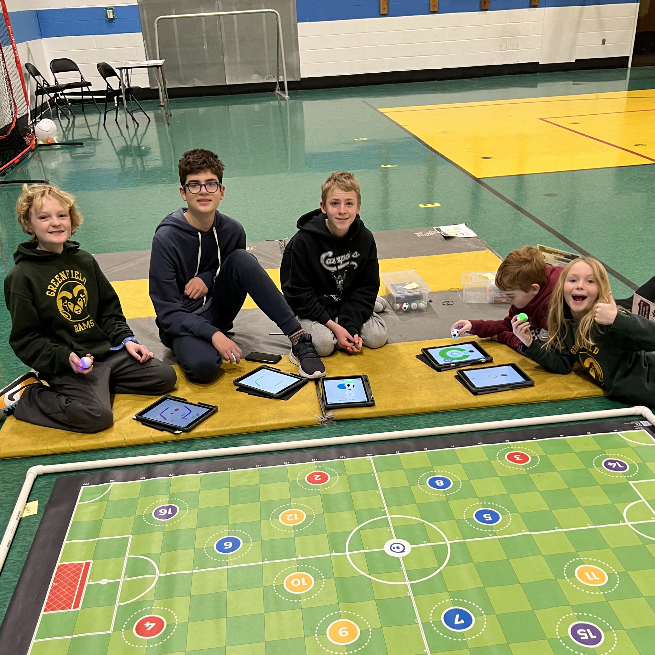 Students sitting on a gym floor at Albert M. Greenfield School using tablets and small devices during a classroom activity